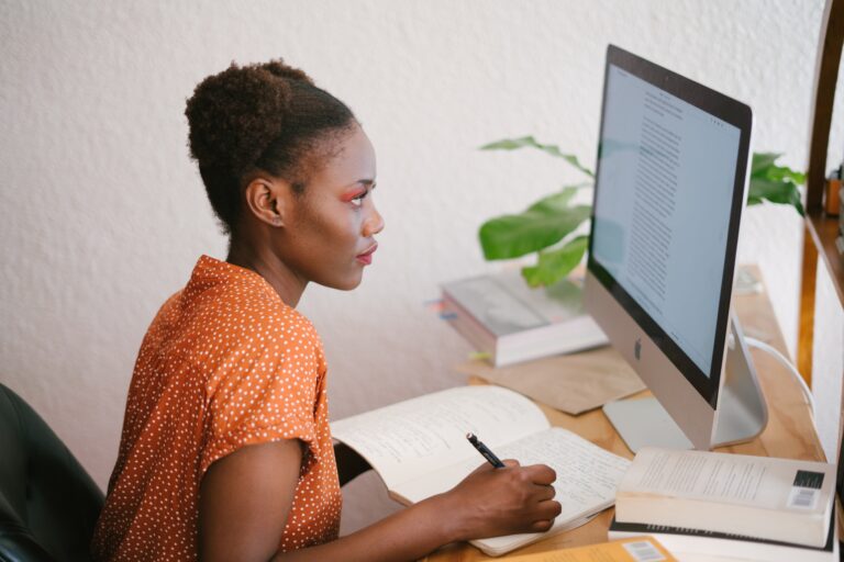 Person at desk looking at computer and writing in notebook